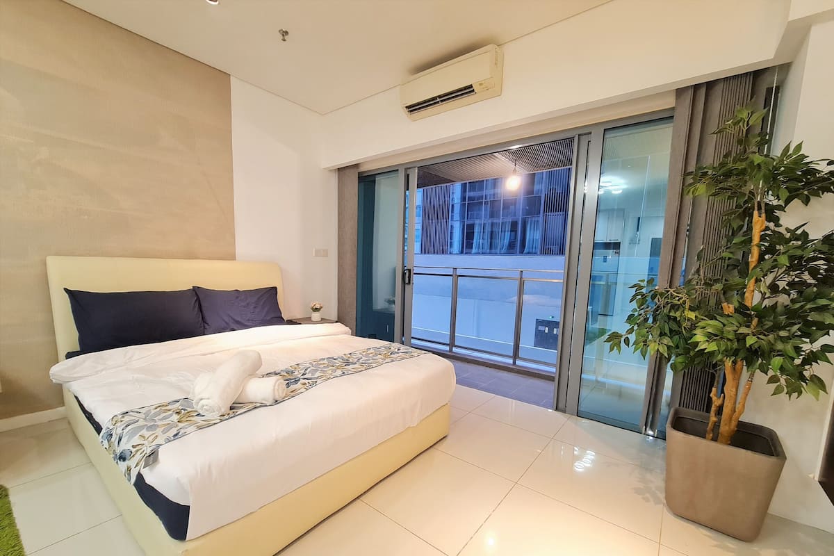 A modern hotel room with a large bed, a potted plant, and a view of the city through the balcony doors.