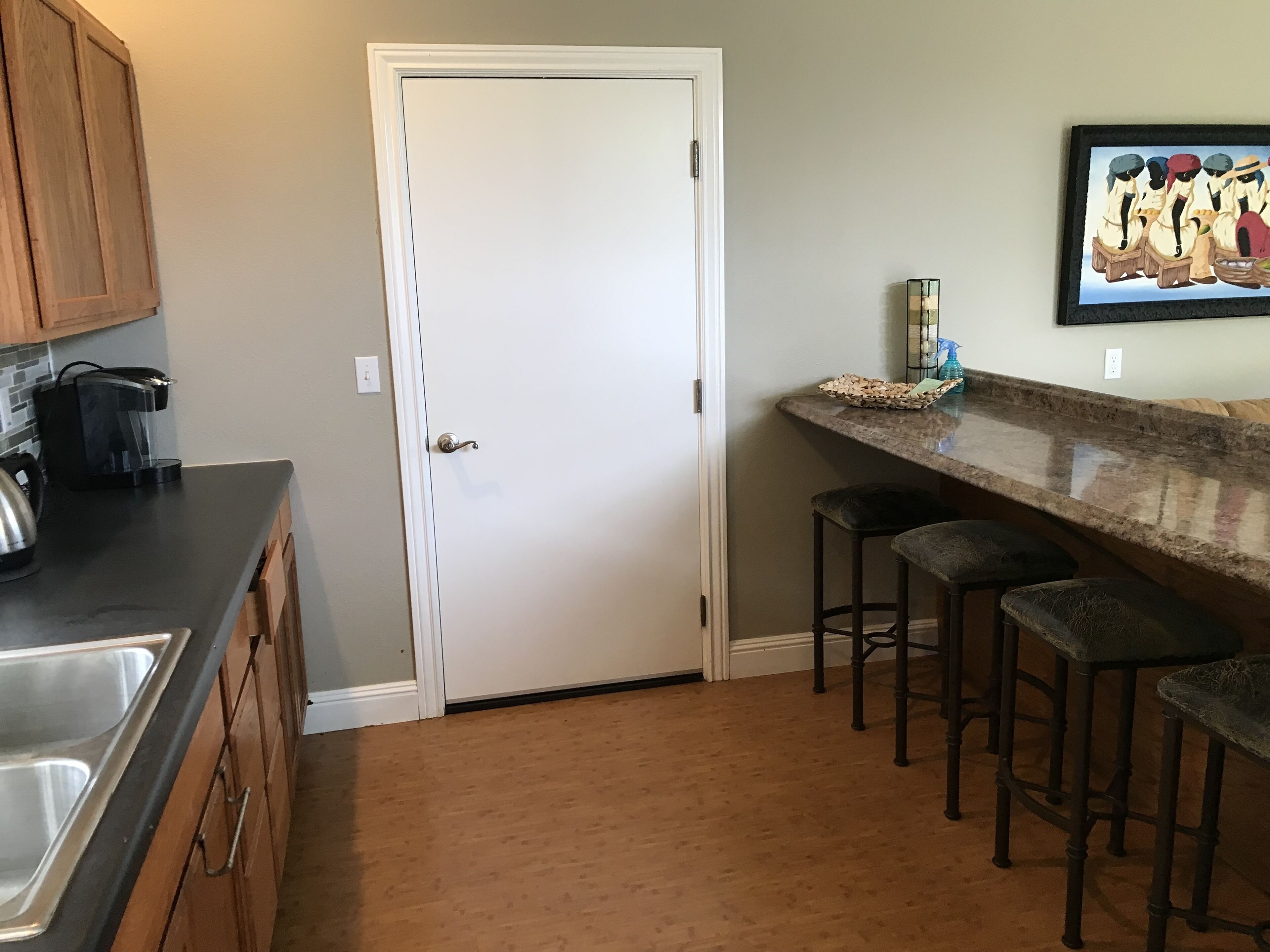 A kitchen with a white door, a coffee maker, a sink, and a bar area with stools.