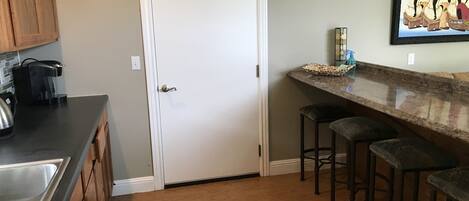 A kitchen with a white door, a coffee maker, a sink, and a bar area with stools.