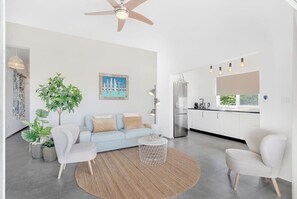 A modern living room with a light blue sofa, white armchairs, and a round coffee table. There is a large plant, a painting on the wall, and a kitchen area visible through an open door. - Belnem Breeze Kas Gosa with private garden (Kralendijk)