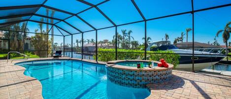 Piscina con zona techada, bañera de hidromasaje y vista a un barco y palmeras.