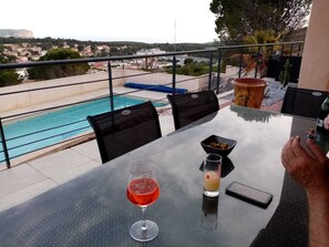 Une table avec un verre de vin rouge, un verre d’une boisson jaune et un téléphone intelligent, le tout sur un balcon donnant sur une piscine et offrant une vue sur la ville.