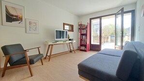 Una sala de estar con un sofá gris, una silla de madera, un escritorio con computadora, una estantería y un ventanal grande.