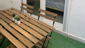 Een houten tafel met stoelen en potplanten, een glazen schuifdeur die leidt naar een slaapkamer met een bed en nachtkastje.