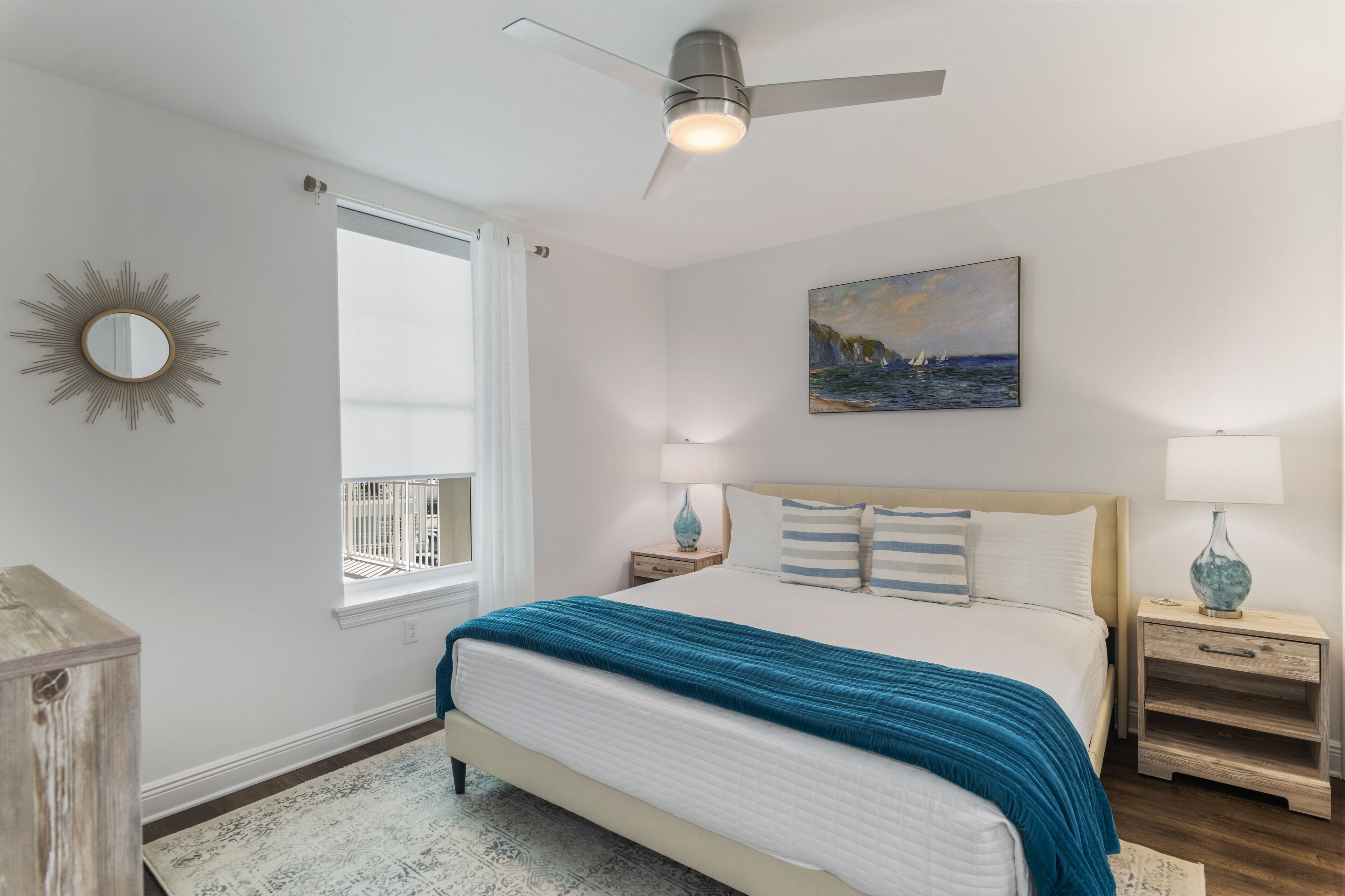 Une chambre à coucher comprenant un lit, des tables de chevet, un ventilateur de plafond, une fenêtre avec des rideaux, un tableau et un miroir décoratif en forme de soleil accroché au mur.