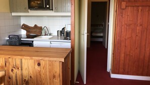 Une cuisine compacte équipée d’un four à micro-ondes, d’un évier et d’un plan de travail en bois. Une porte donne accès à une chambre à coucher qui comprend un lit et une moquette rouge.