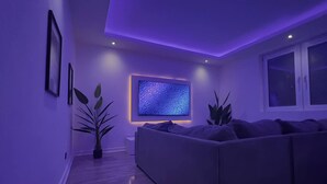 Una sala de estar moderna con un televisor de pantalla plana grande montado en la pared, un sofá gris y dos plantas en macetas.