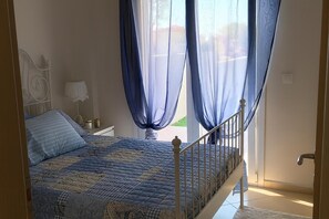 A bedroom with a bed, a nightstand, a lamp, and sheer blue curtains. - Palm Garden Apartment (Sithonia)