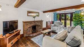 A living room with a brick fireplace, a television on a wooden stand, a grey sofa, and a view through large glass doors to an outdoor area. - Church Walk Farm (Snarestone)