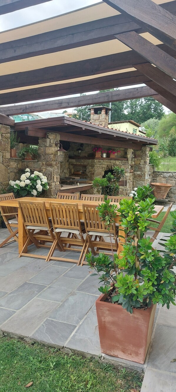 An outdoor patio with stone flooring, wooden tables and chairs, and a stone fireplace.