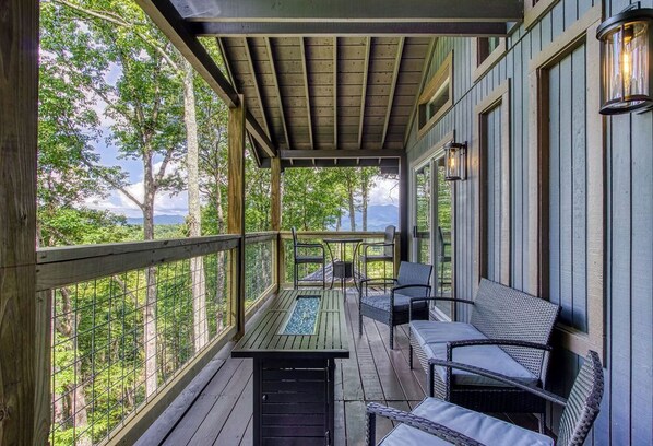 A covered porch with a wooden table and chairs, a view of trees and mountains, and a railing.