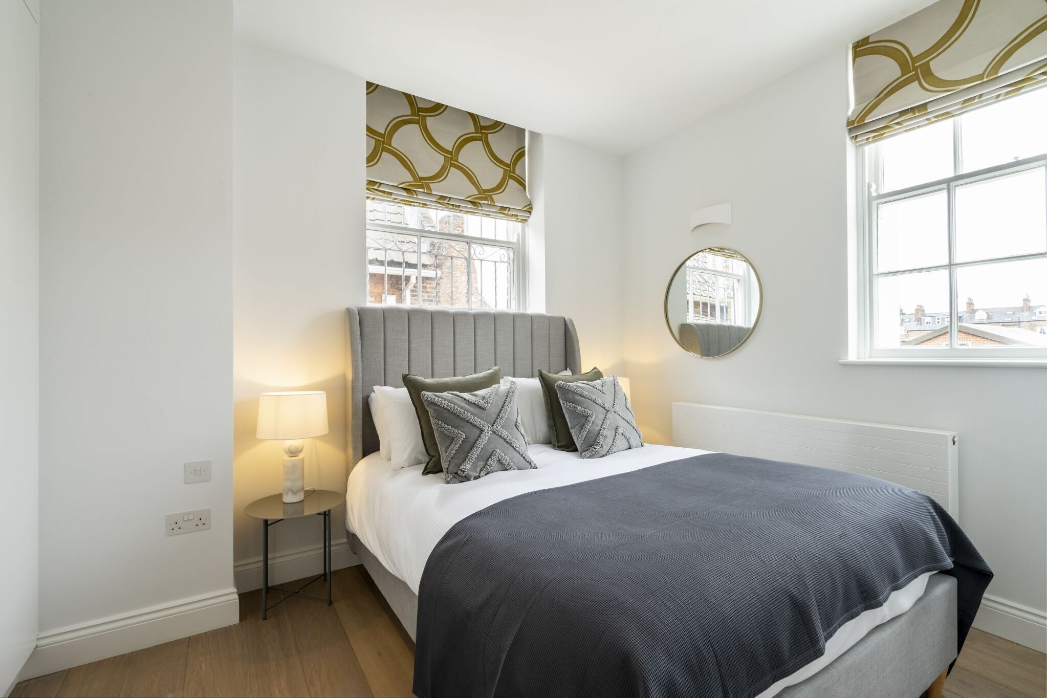 A bedroom with a bed, bedside table, lamp, window with blinds, and a mirror on the wall.