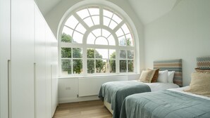 Een slaapkamer met een groot rondboogvenster, twee bedden en hoofdeinden met een patroon.