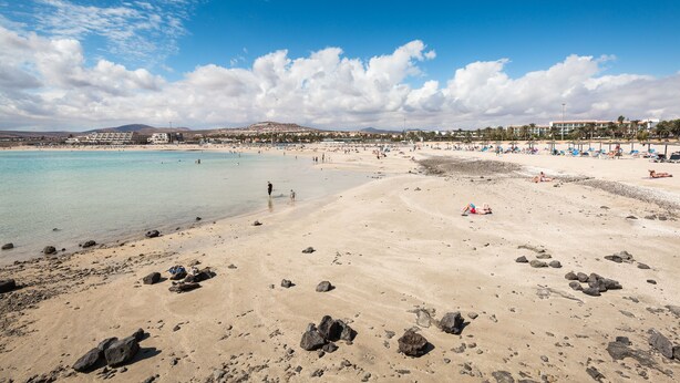 Ein Strand mit klarem türkisblauem Wasser, felsigem Ufer und einem Resorthintergrund mit Gebäuden.