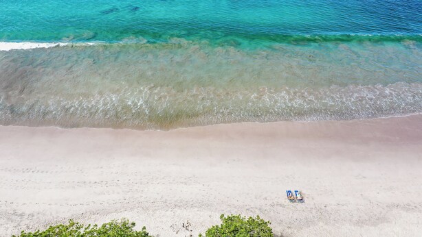 Une vue aérienne d’une plage aux eaux turquoise et limpides, avec un rivage sablonneux et une seule chaise de plage.