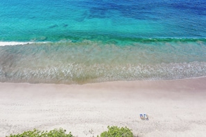 Eine Vogelperspektive zeigt einen Strand mit klarem türkisblauem Wasser, einem sandigen Ufer und einem einzelnen Liegestuhl.
