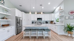 A modern kitchen with white cabinets, a stainless steel refrigerator, a central island with bar stools, pendant lights, and a microwave.