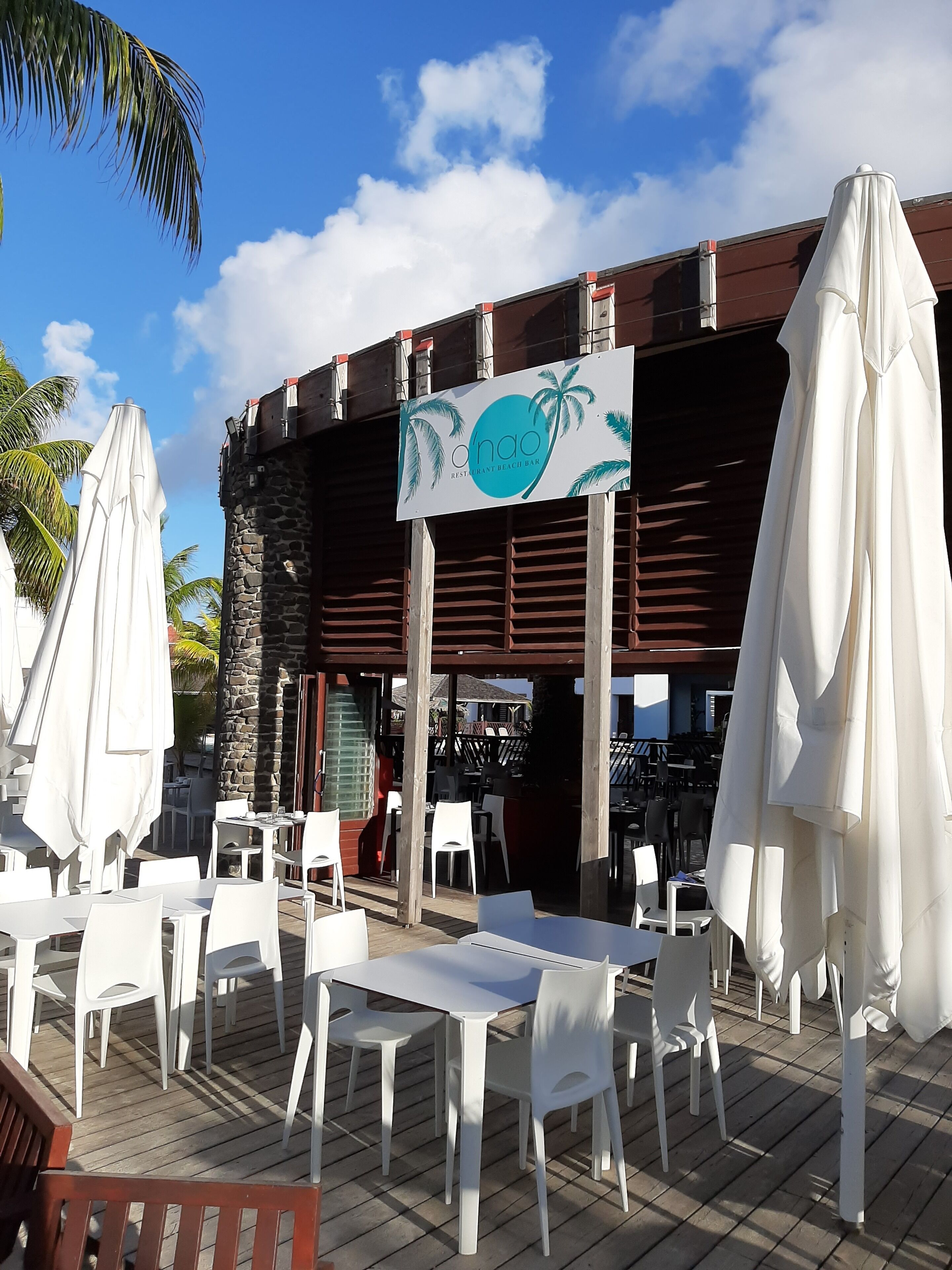 Uma área de estar externa com cadeiras e mesas de plástico branco, uma placa indicando 'O'NaO Restaurant Beach Bar' e palmeiras ao fundo.