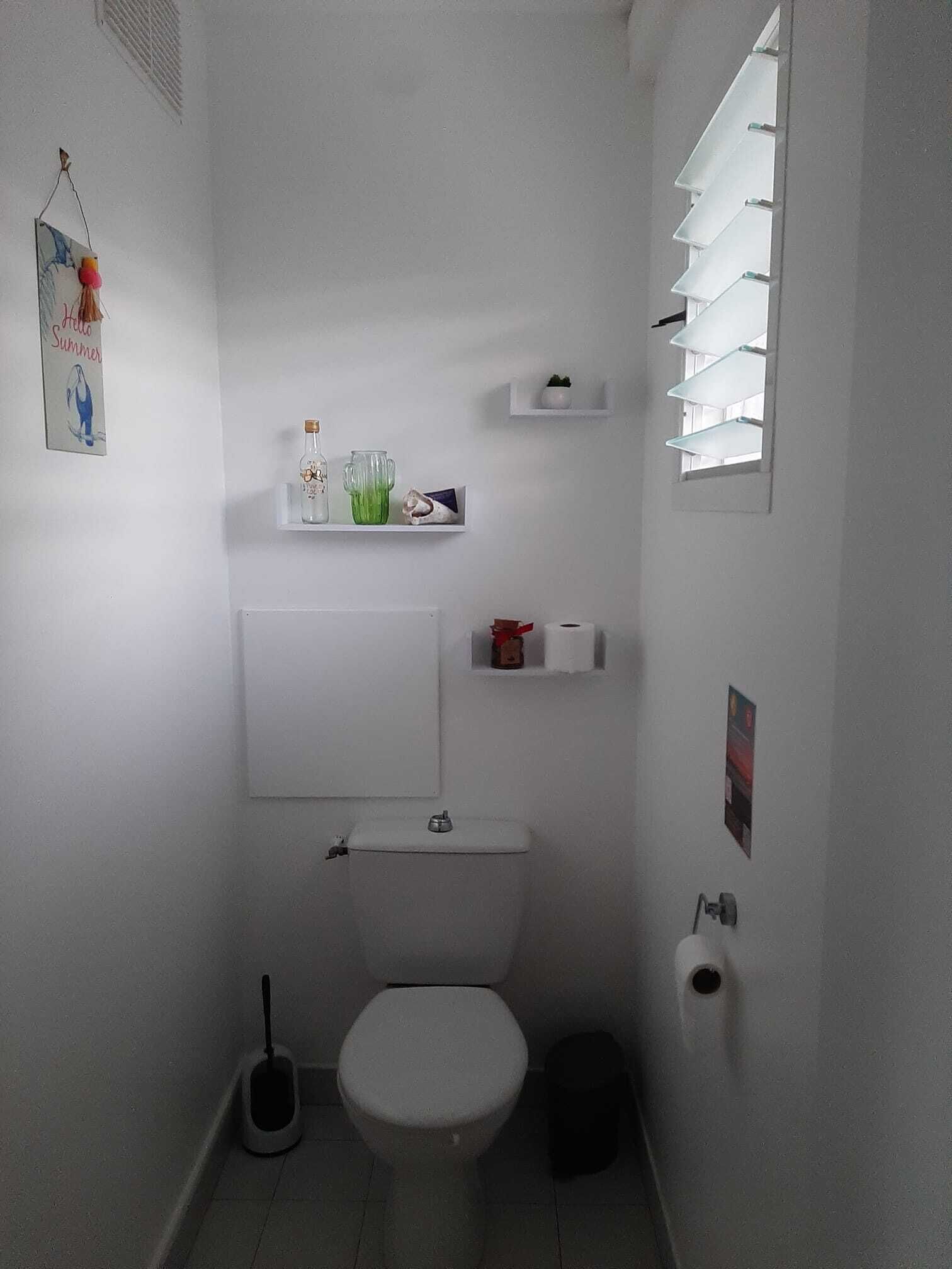 Een kleine, witte badkamer met een toilet, een hygiëneschuimer en een plank met toiletartikelen.