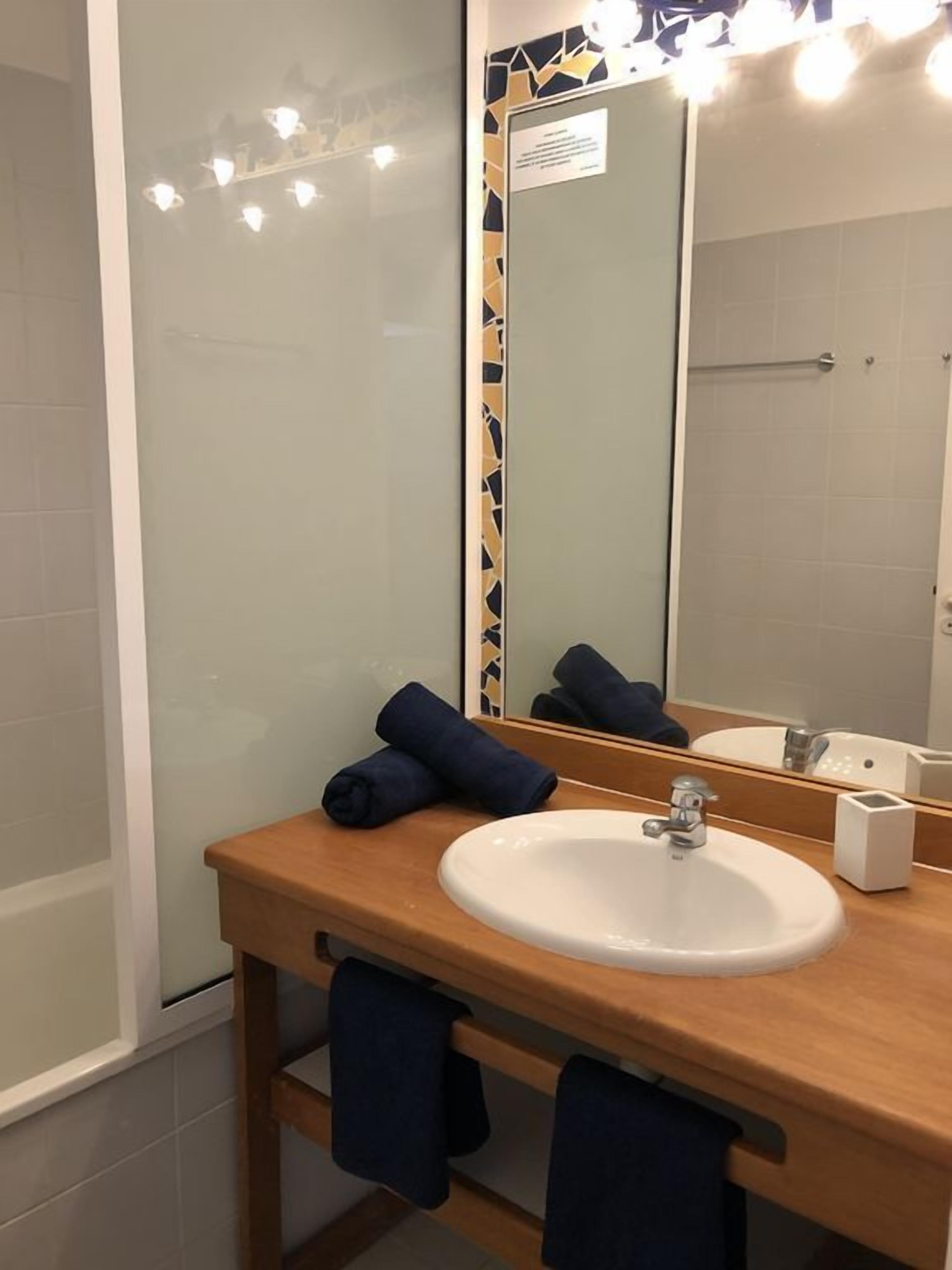 Un baño con tocador de madera, un lavabo blanco y un espejo grande. Hay toallas en la encimera y se ve una zona de ducha al fondo.