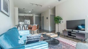 Ein modernes Wohnzimmer mit einem blauen Sofa, einem Couchtisch und einem Flachbildfernseher. Im Hintergrund befindet sich eine Essecke mit Stühlen und einem Tisch.