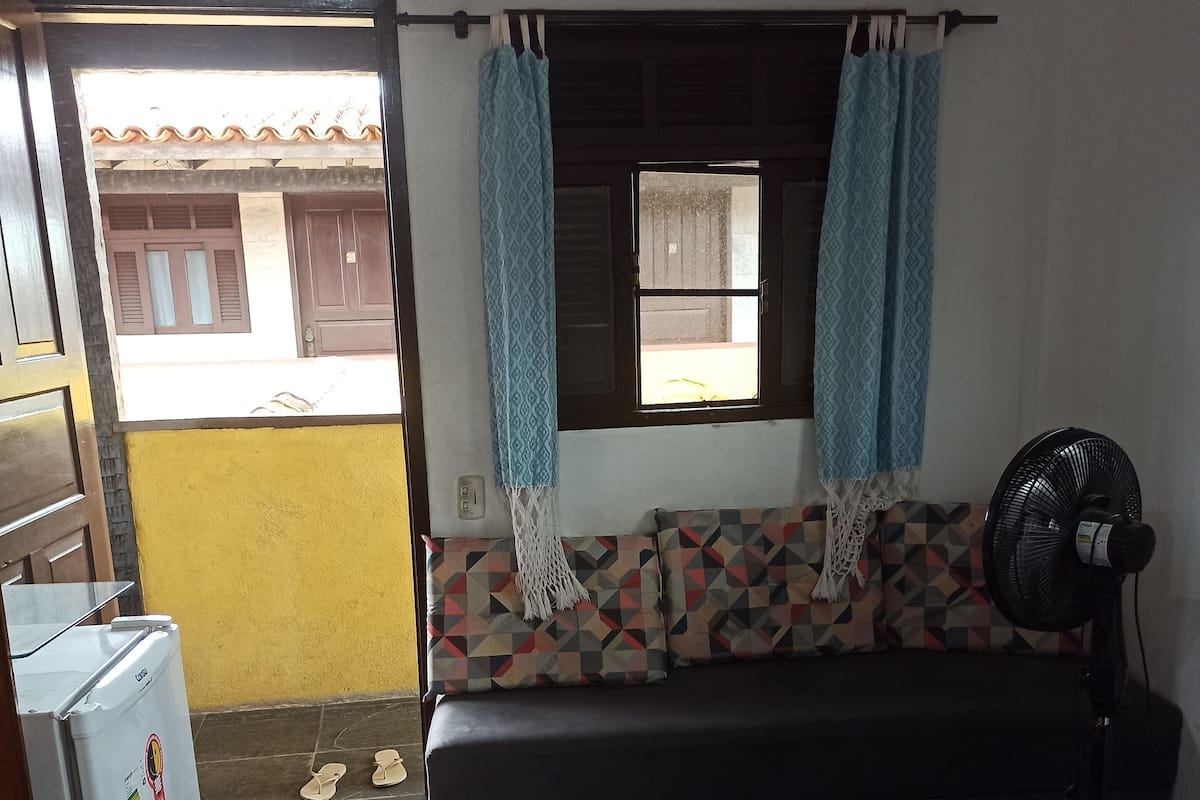 Une pièce de taille modeste, comprenant un canapé, une petite table, un ventilateur et un réfrigérateur.