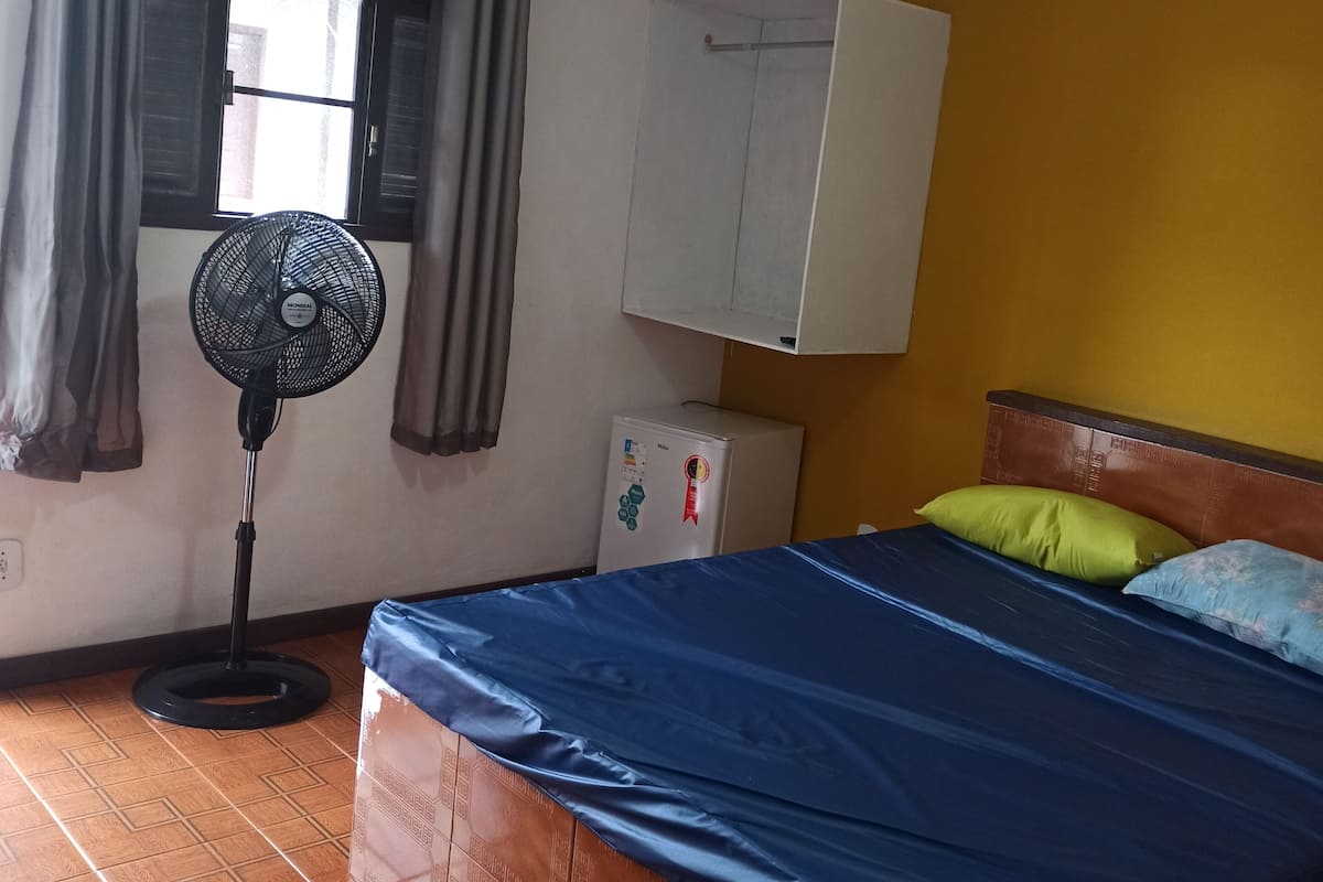 Une chambre avec un lit, un ventilateur sur pied, un petit réfrigérateur et une fenêtre avec des rideaux.
