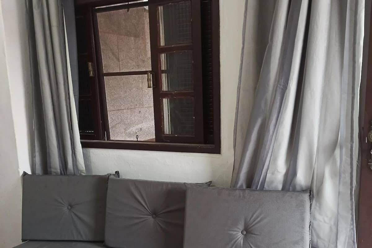 Un canapé gris avec trois coussins, une fenêtre avec des stores et des rideaux gris.