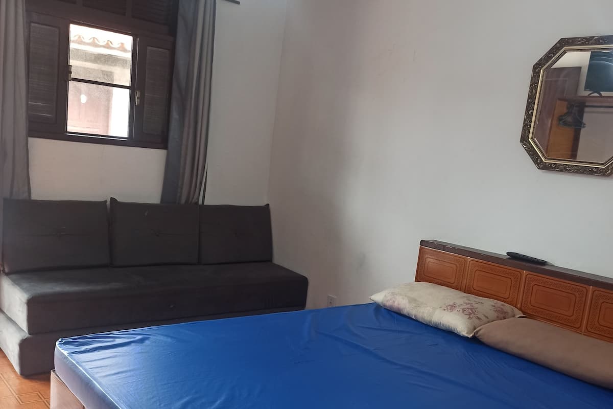 Une chambre à coucher avec un lit, un canapé, une fenêtre avec des rideaux, un ventilateur de plafond et un miroir au mur.