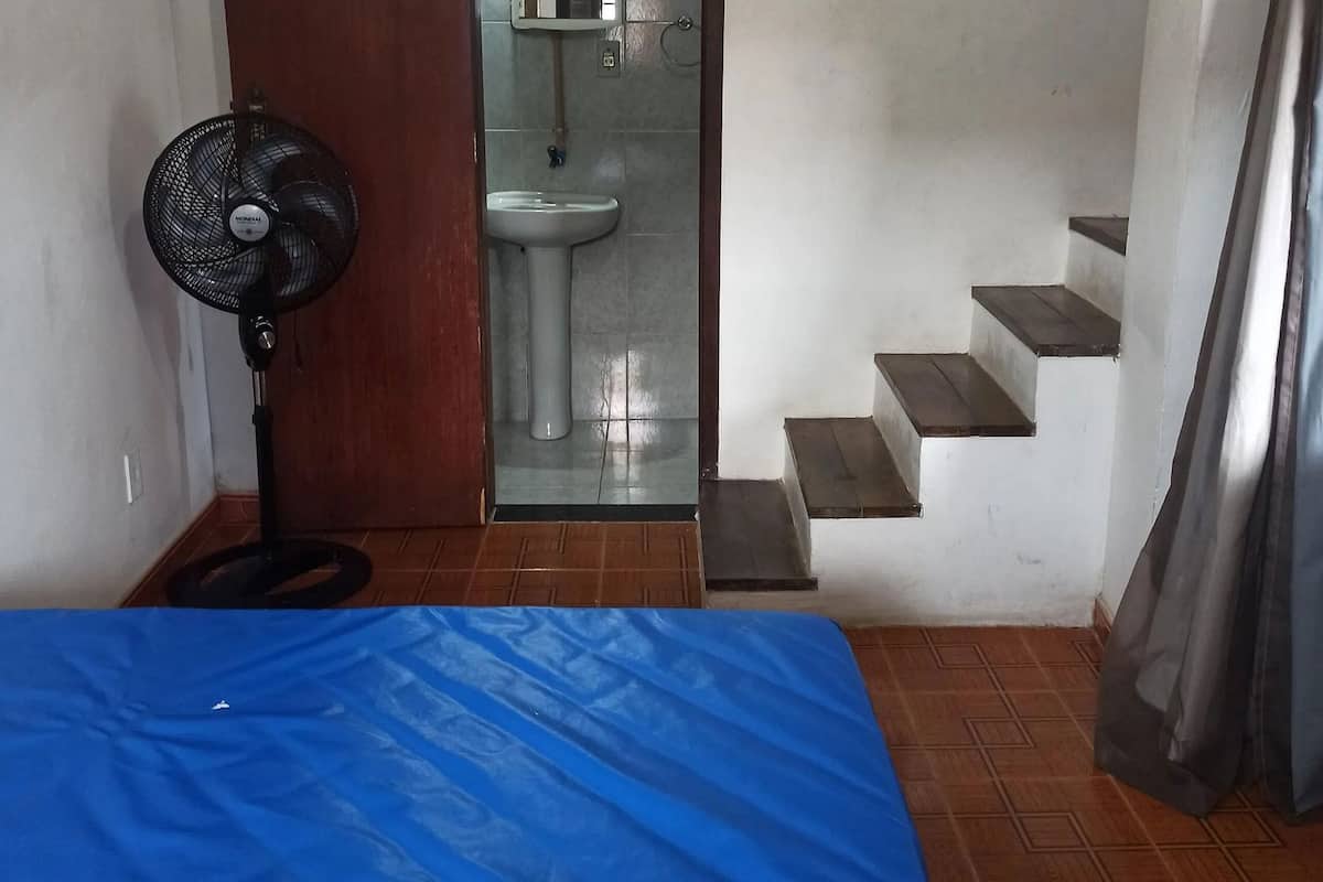 Une pièce comprenant un lit, un ventilateur sur pied, un lavabo et un escalier.