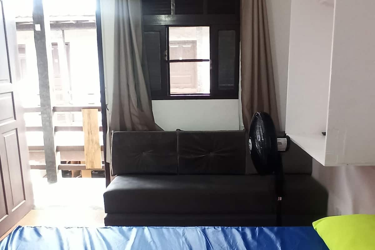 Une pièce avec un lit, une fenêtre avec des rideaux, un ventilateur et un tableau blanc.