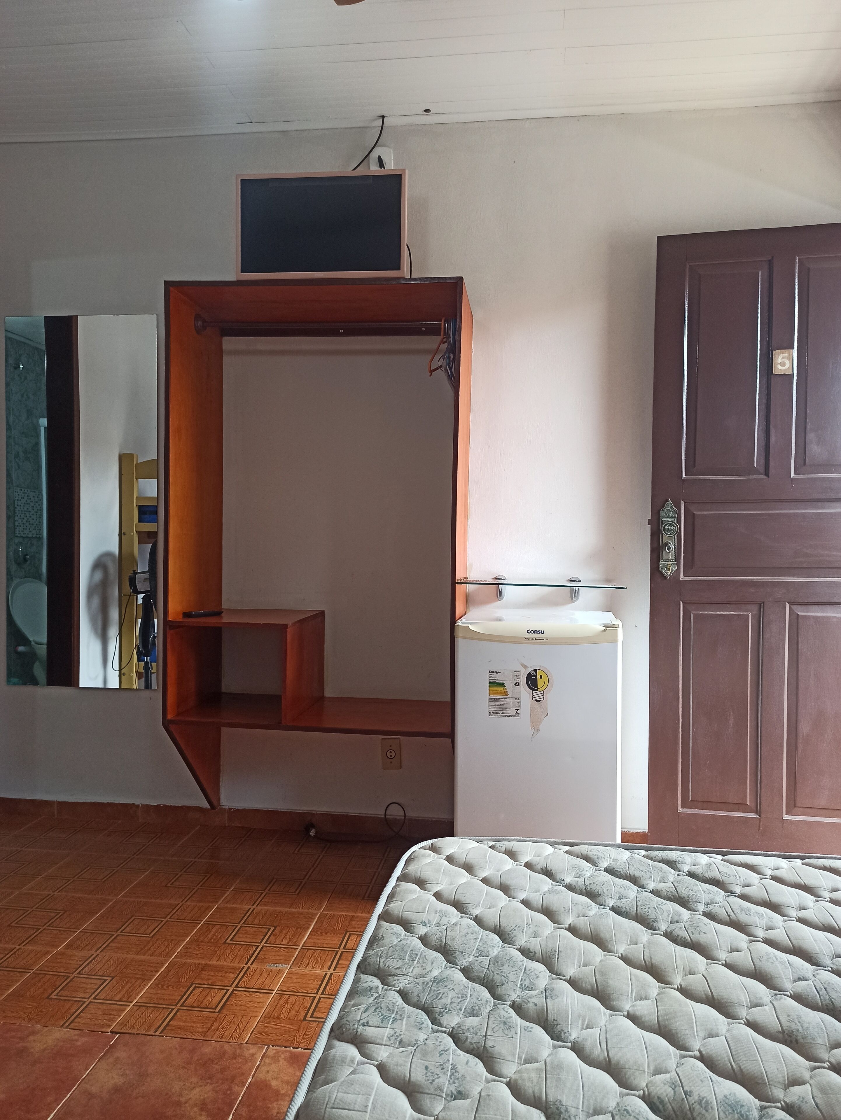 Una habitación con una cama, un estante de madera, un refrigerador blanco y una puerta marrón.