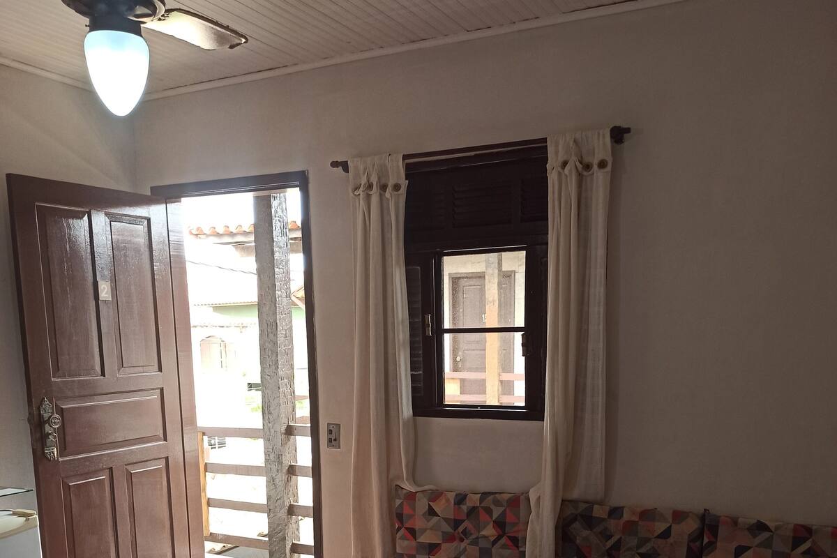 Une pièce avec un lit, une fenêtre avec des rideaux, un ventilateur de plafond et une porte donnant sur l’extérieur.
