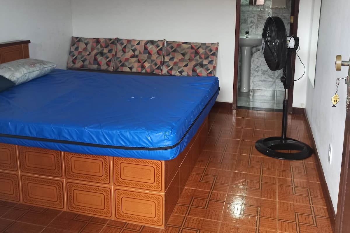 Une chambre à coucher avec un lit, un ventilateur de plafond, un ventilateur sur pied et une salle de bain visible à travers une porte ouverte.