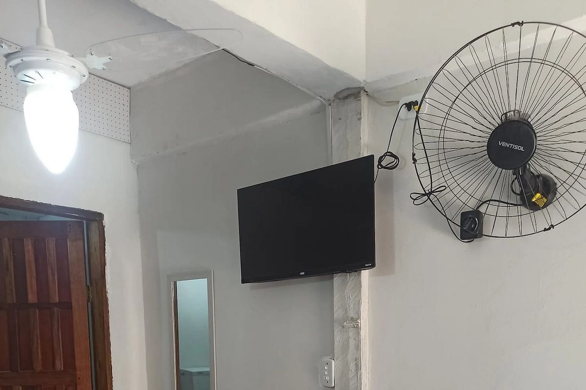 Une pièce équipée d’un téléviseur fixé au mur, d’un ventilateur sur pied, d’un luminaire au plafond et d’une porte.