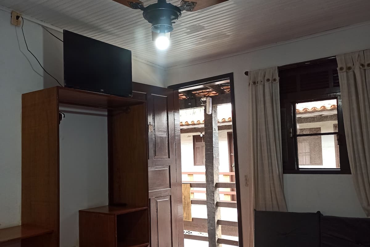 Une chambre avec un lit, une armoire, un ventilateur de plafond, un téléviseur posé sur l’armoire et une fenêtre avec des rideaux.