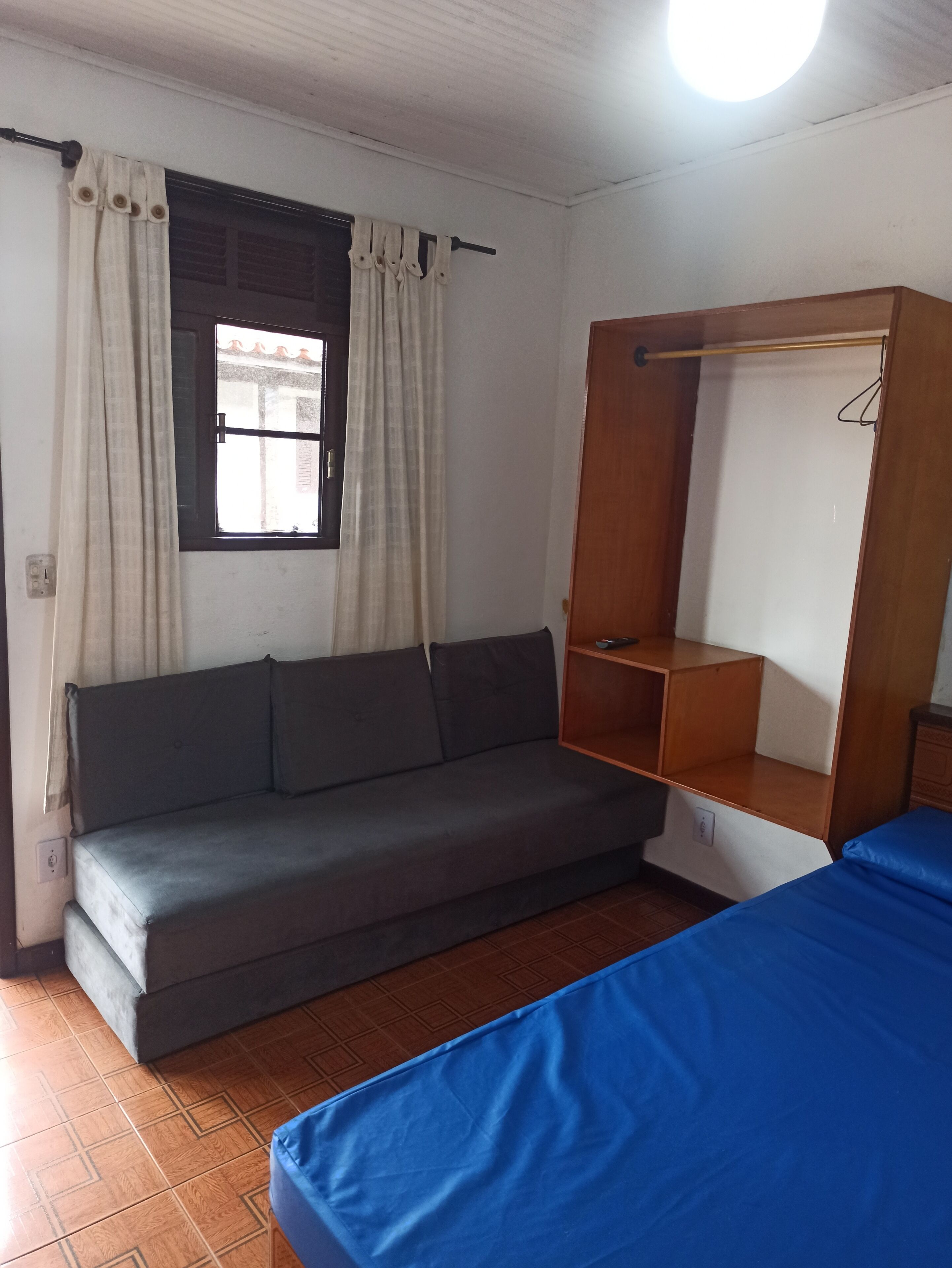 Una habitación con un sofá gris, una cama azul, un armario de madera y una ventana con cortinas.