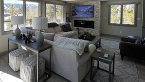 A modern living room with a large sofa, a coffee table, ottomans, a TV mounted on a stone wall, and large windows offering a view of trees and mountains. -  Walk To Town, Ski In Style - Stunning Views w/Hot Tub, 1 block to Free Shuttle (Breckenridge)