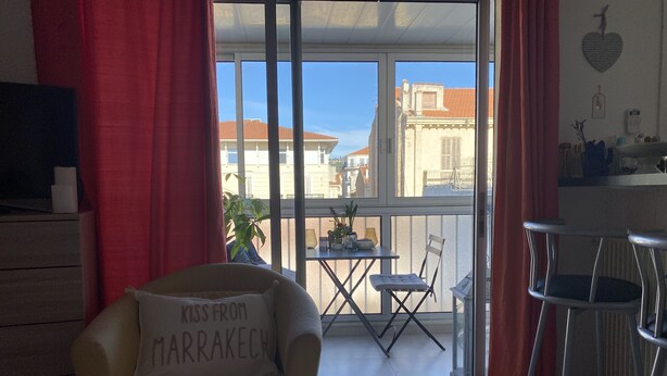 Ein gemütliches Zimmer mit einem Bett, einem Sessel mit Kissen, einem kleinen Tisch und einem Balkon mit Blick auf Gebäude.