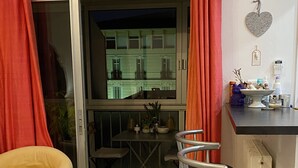 Ein gemütlicher Wohnraum mit einem gelben Sessel, einem Esstisch und zwei Barhocker. Eine Glastür öffnet sich zu einem Balkon mit Blick auf Gebäude.