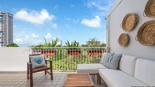 A balcony with a white sofa, wooden table, and wicker baskets. - Beautiful Queen Penthouse with Large Terrace (San Juan)