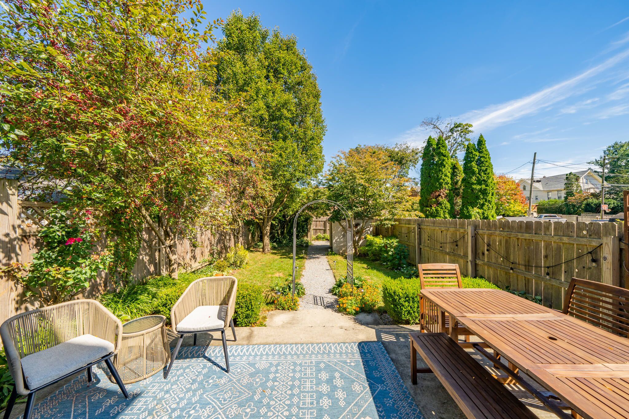 Un jardin avec une table et des chaises en bois, un tapis à motifs et un chemin menant à une maison.
