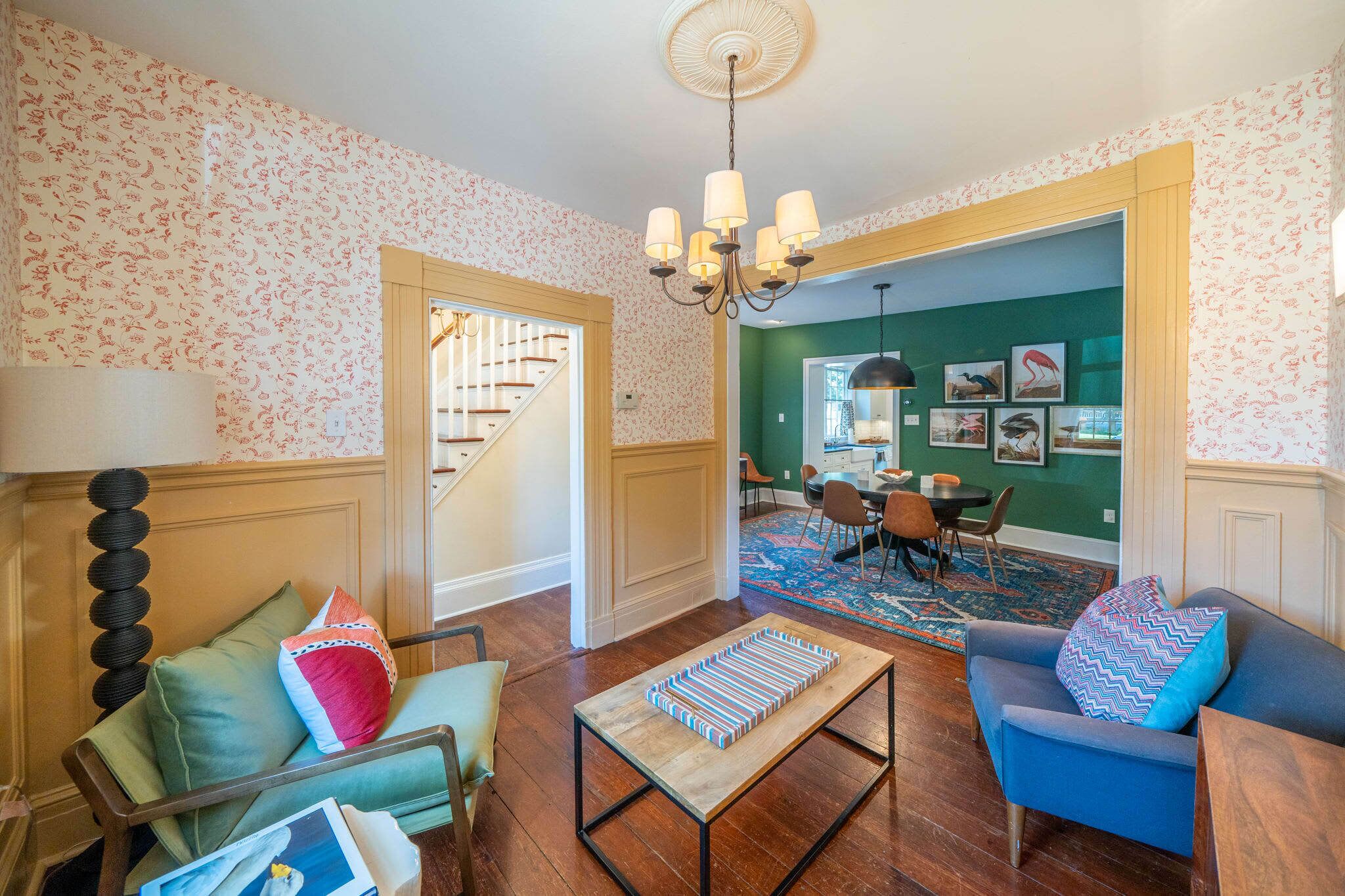 Una sala de estar con una mesa de centro de madera, un sofá azul y un sillón verde. Se ve una escalera que conduce a otra habitación, y un comedor con sillas y una mesa.