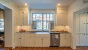 A kitchen with white cabinets, a herringbone-patterned floor, a window with curtains, and a stainless steel dishwasher. - Spacious 3BR Historic Home | Walk Downtown+Parking (Kennett Square)