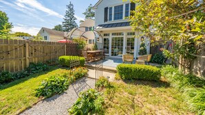A house with a patio, a wooden fence, and a garden with various plants and a small table with chairs. - Spacious 3BR Historic Home | Walk Downtown+Parking (Kennett Square)