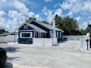 Una casa de una sola planta con fachada azul, una cerca blanca y un camino de acceso a la puerta principal.