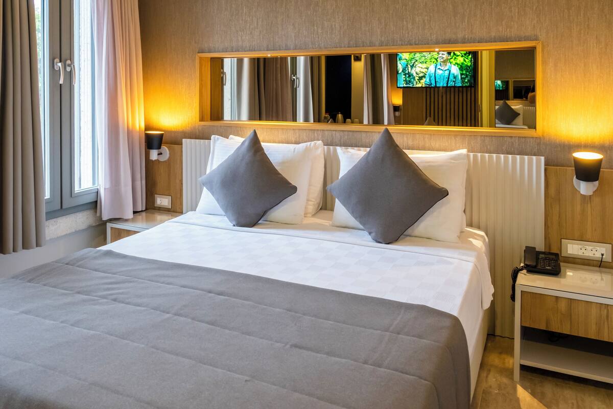 A hotel room with a large bed, two bedside tables, a bedside lamp, a flat-screen TV mounted on the wall, and a window with curtains.
