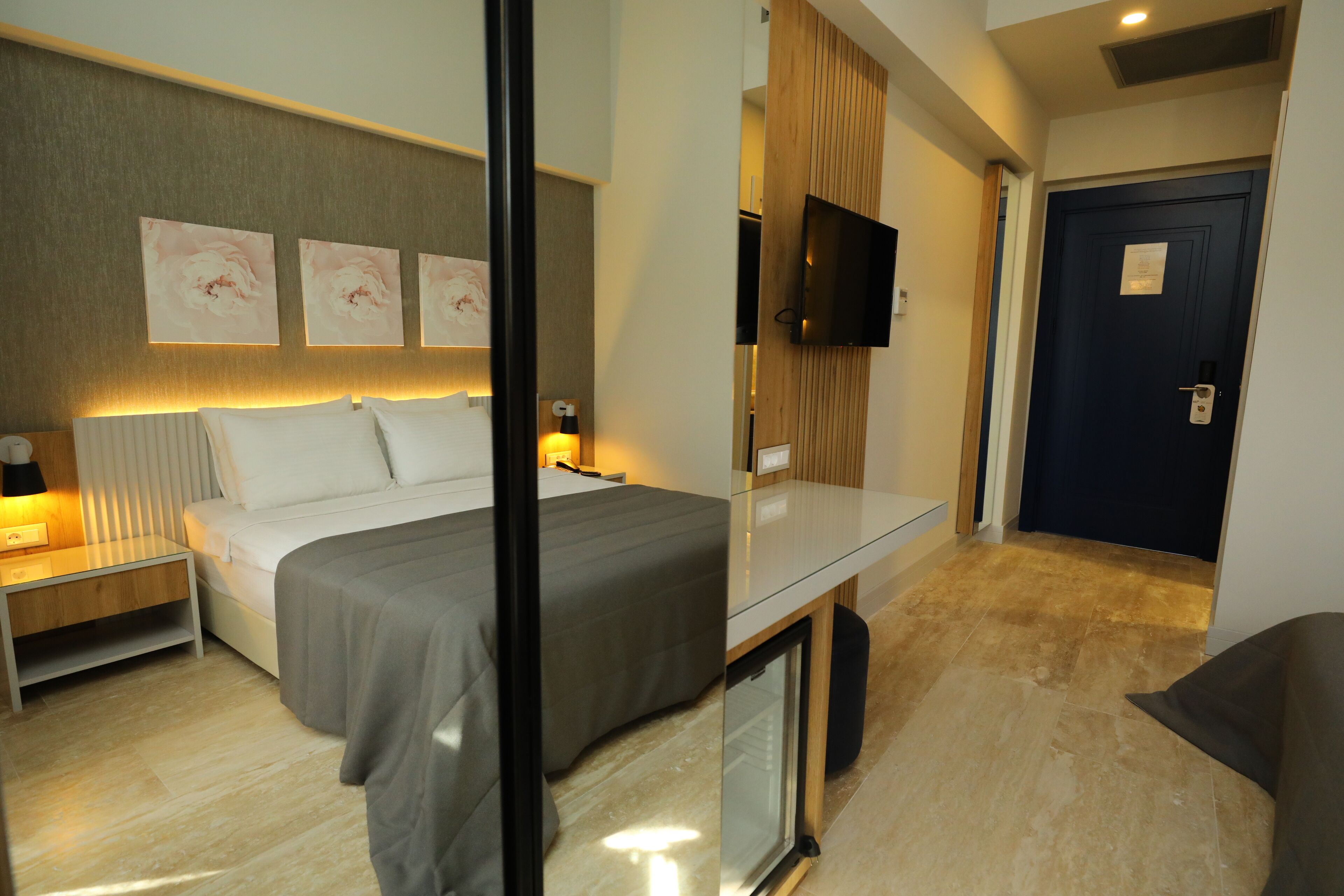 A modern hotel room with a bed, bedside tables, a TV, and a door.