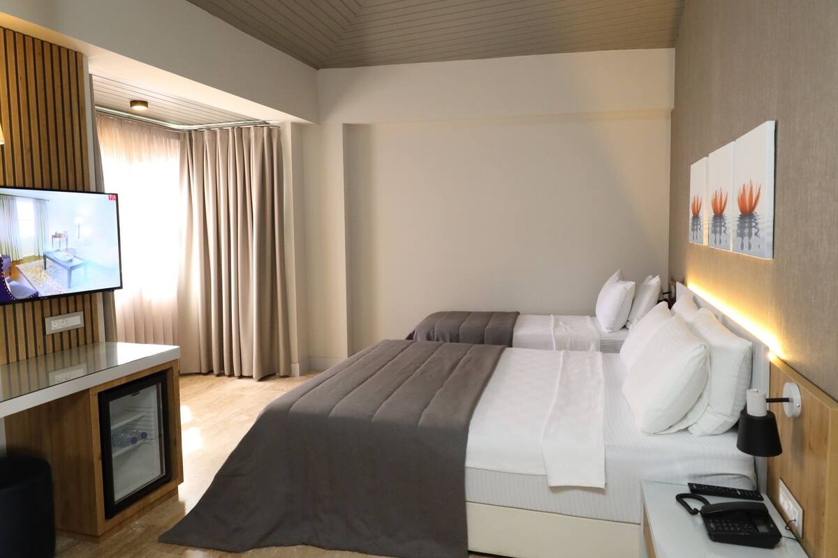 A modern hotel room with a large bed, a bedside table with a lamp, a television, and a small desk with a computer.