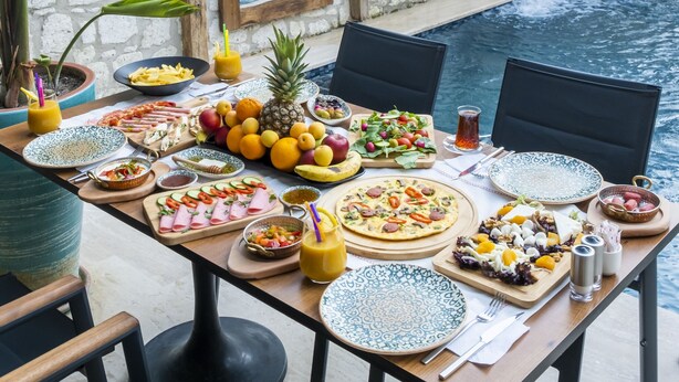 A dining table set with various dishes and drinks, surrounded by chairs, with a pool and plants in the background. - Ariel Hotel Alaçatı (Cesme)